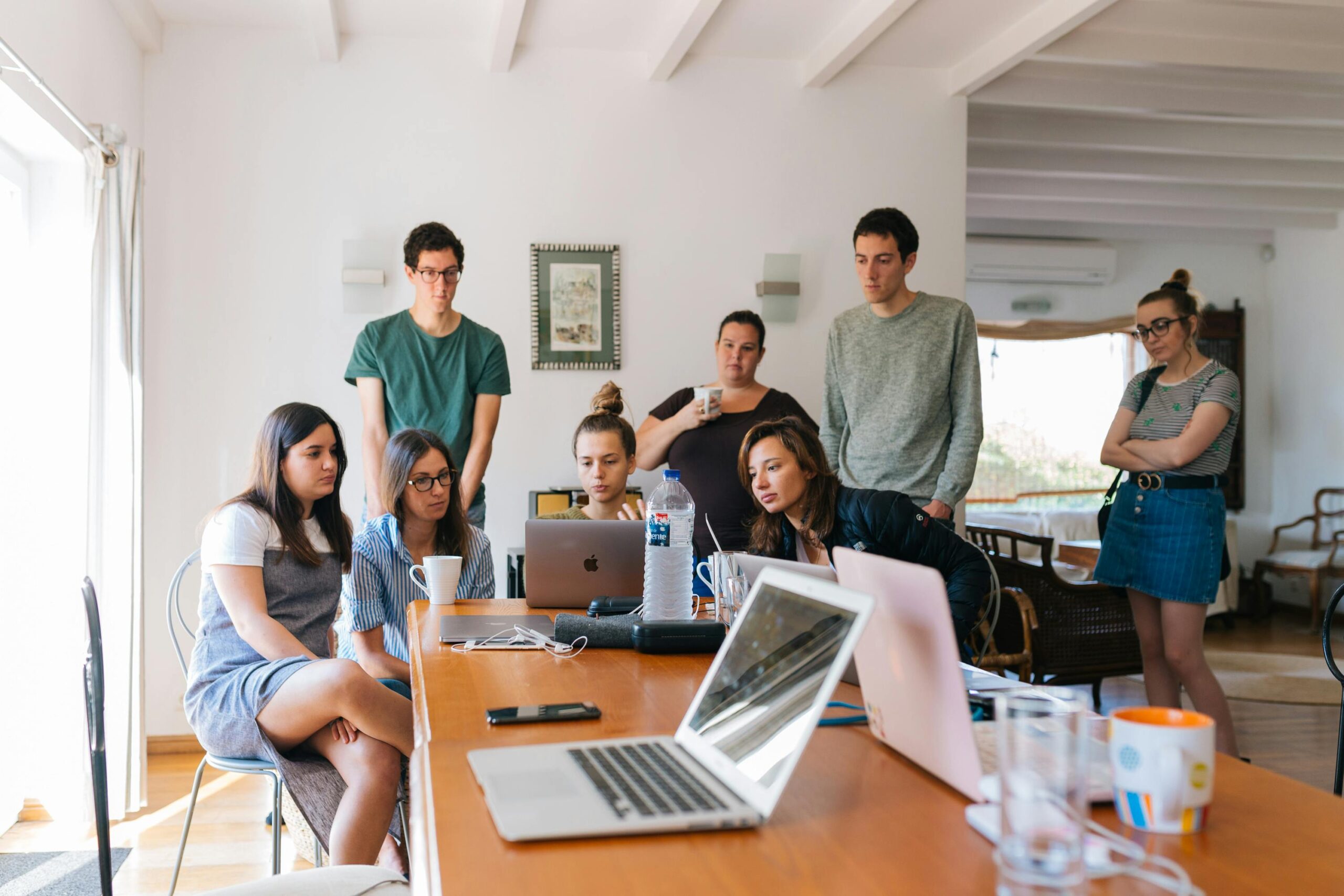 A team of 8 marketing professionals are sitting in a meeting. They are looking towards the same laptop screen discussing their company website. 