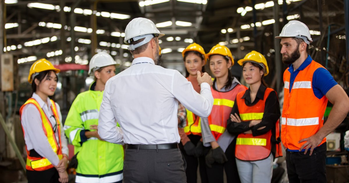 diverse group of factory workers in hard hats and safety vests listening to a supervisor during a training session on the production floor