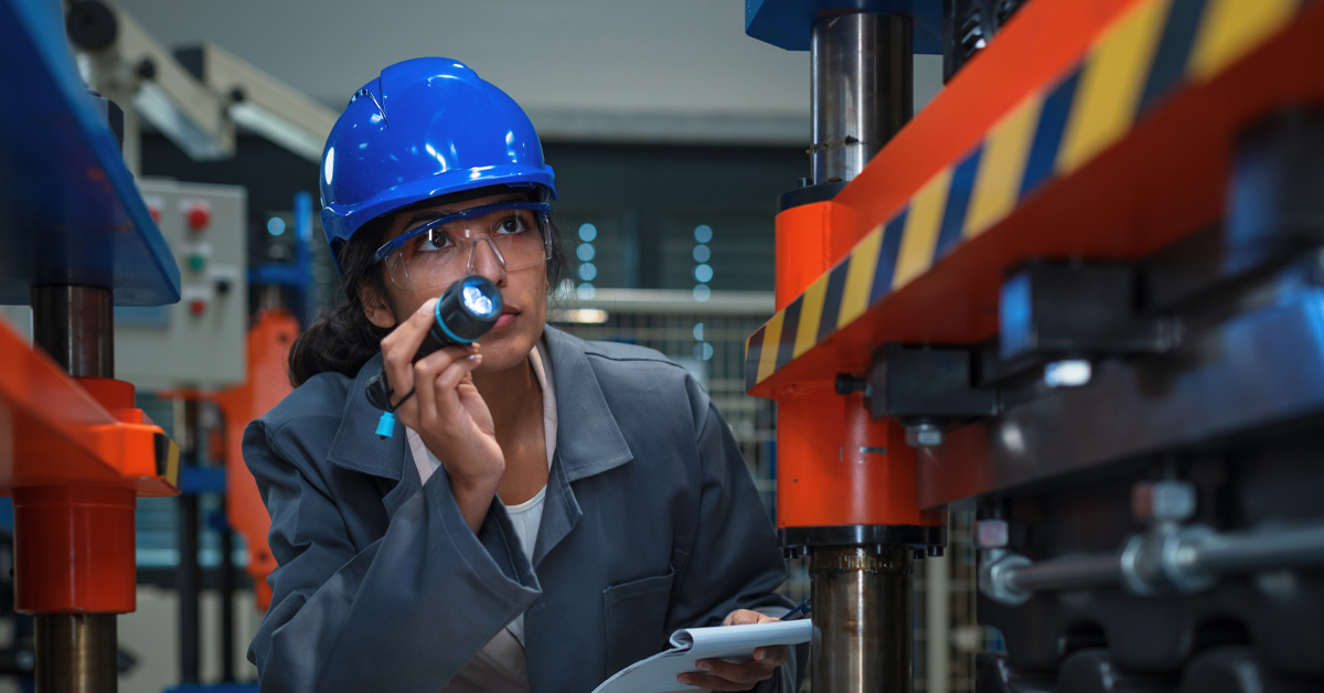 A technician wearing a blue hard hat and safety glasses uses a flashlight to perform a detailed safety inspection on industrial machinery
