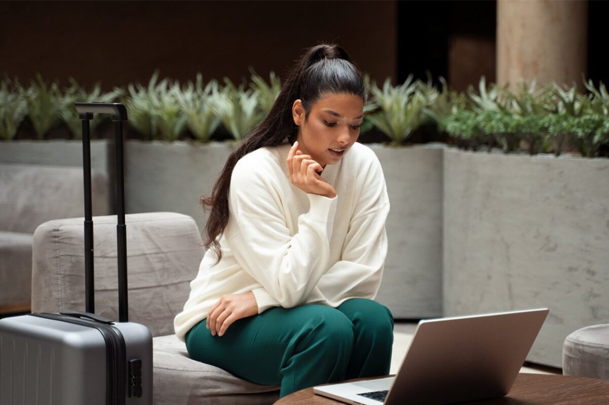 A woman in a white sweater and green trousers looks at a laptop on a coffee table in a modern lounge area. A silver suitcase sits beside her, symbolizing the flexibility of modern travel demand trends.