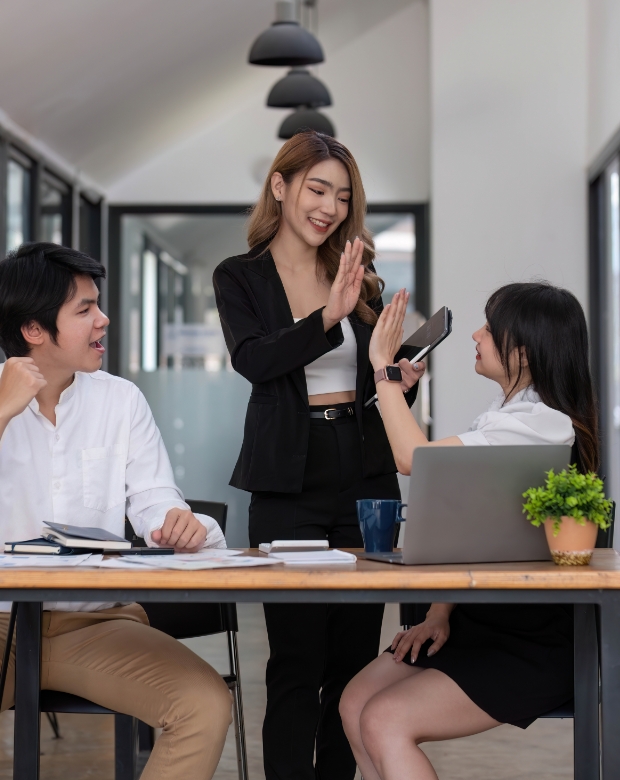 Three young Asian business professionals smiling and giving a high-five during a team meeting with a laptop, representing Traditional Chinese translation and business services