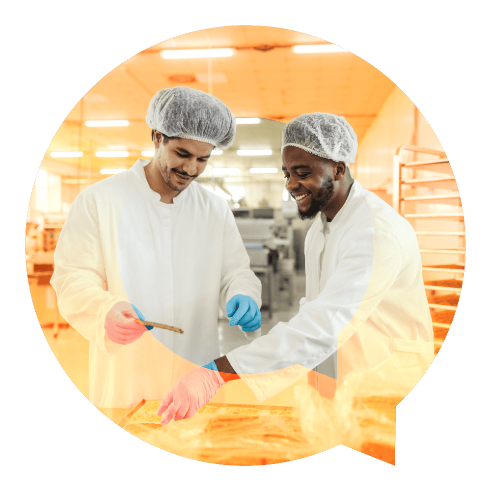 Two food production workers in white coats and hairnets smiling while reviewing work together in a food manufacturing facility
