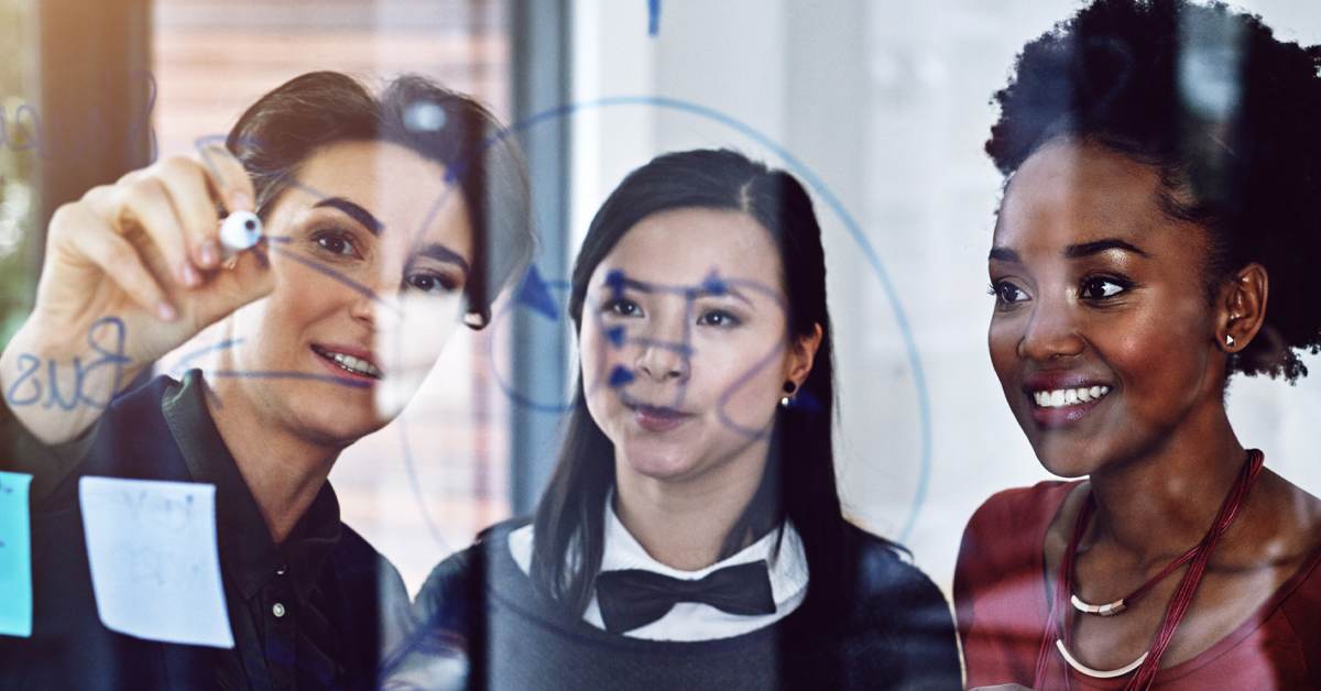Three women collaborating and writing on a glass board in an office, representing insight teams working together on market research strategy