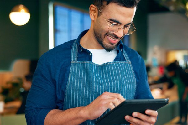 A food service professional in an apron smiling while reviewing training content on a tablet in a cafe environment