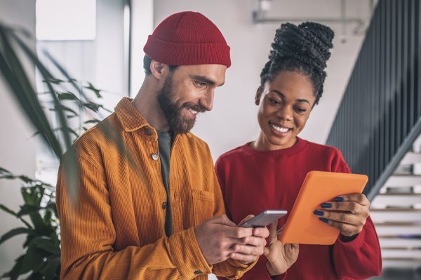 Two creative professionals smiling while reviewing campaign content together on a tablet and phone in a modern office setting
